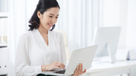 Beautiful young Asian woman in elegant outfit holding and using laptop with smile in modern office